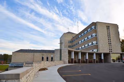Exterior view of Windsor Park senior living facility with driveway, entrance, and cross sculpture.