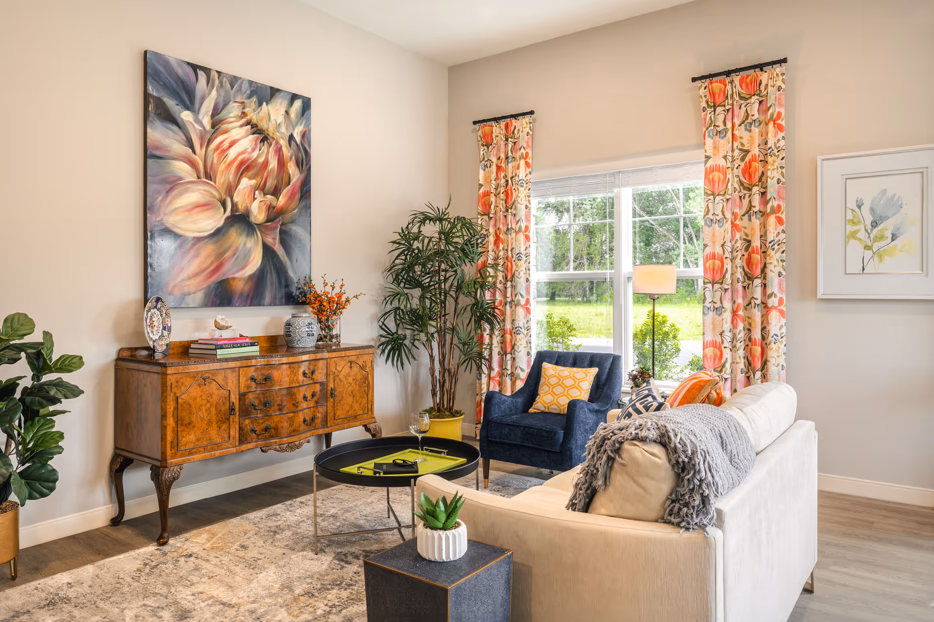 Bright, furnished living room with a sofa, armchair, large window with patterned curtains, wooden sideboard and wall art.