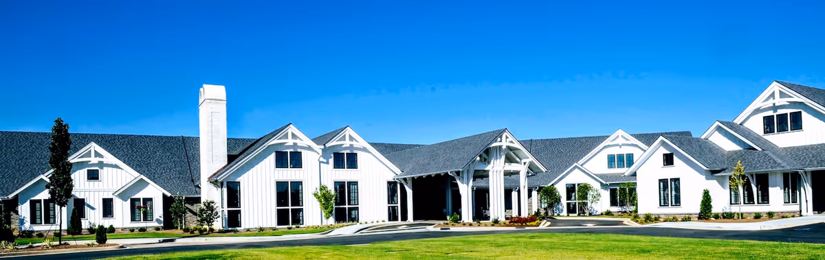 Front view of a long white ranch-style senior living building with multiple gables, a covered entrance, and a manicured lawn under a clear blue sky.