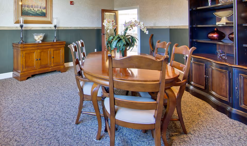 Wooden dining table with chairs and a floral centerpiece in a carpeted communal dining room with cabinetry and decorative accents.