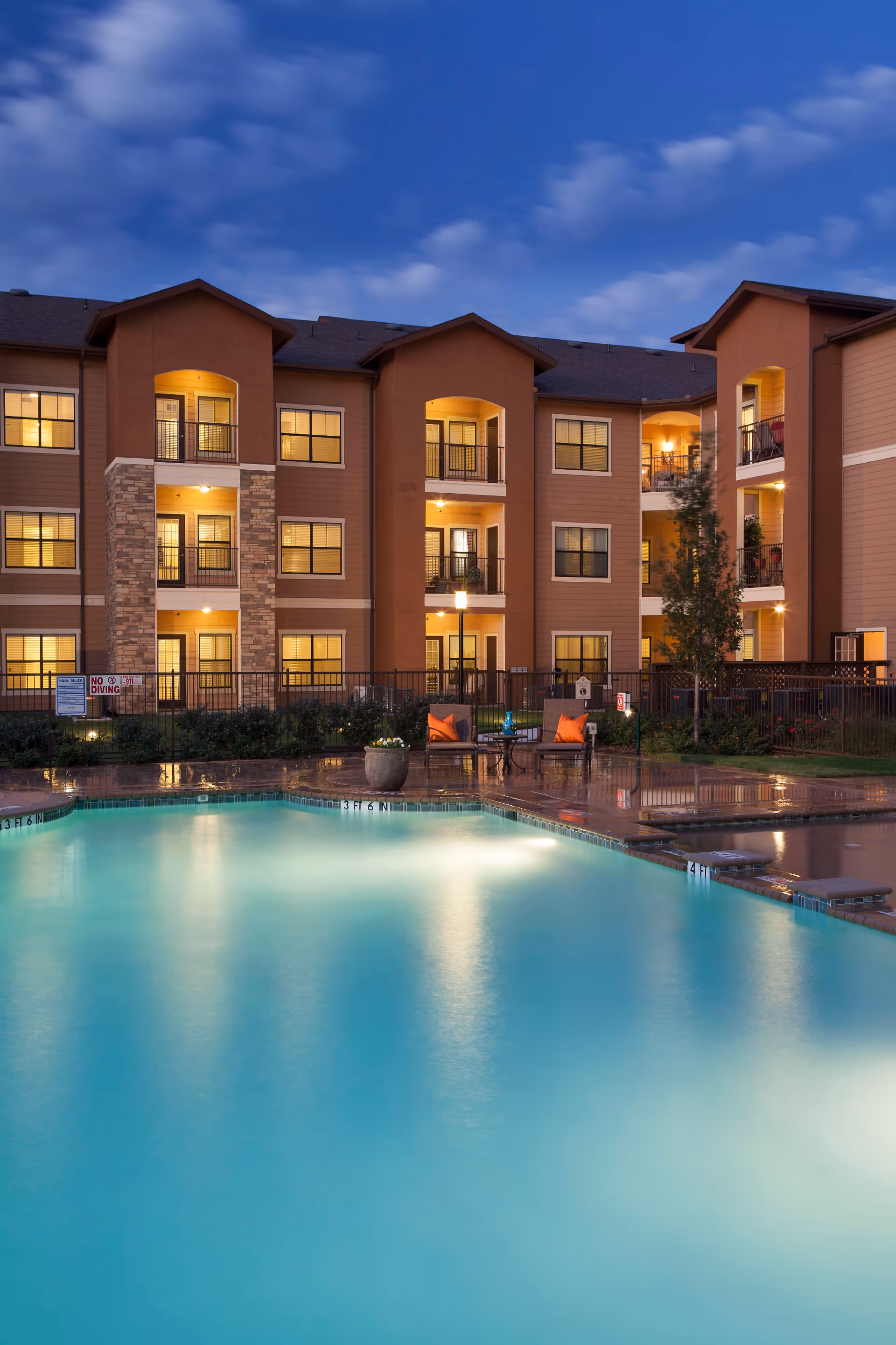 Evening view of a residential building with multiple balconies and windows illuminated by warm lights, overlooking a swimming pool with clear blue water and poolside seating with orange cushions.