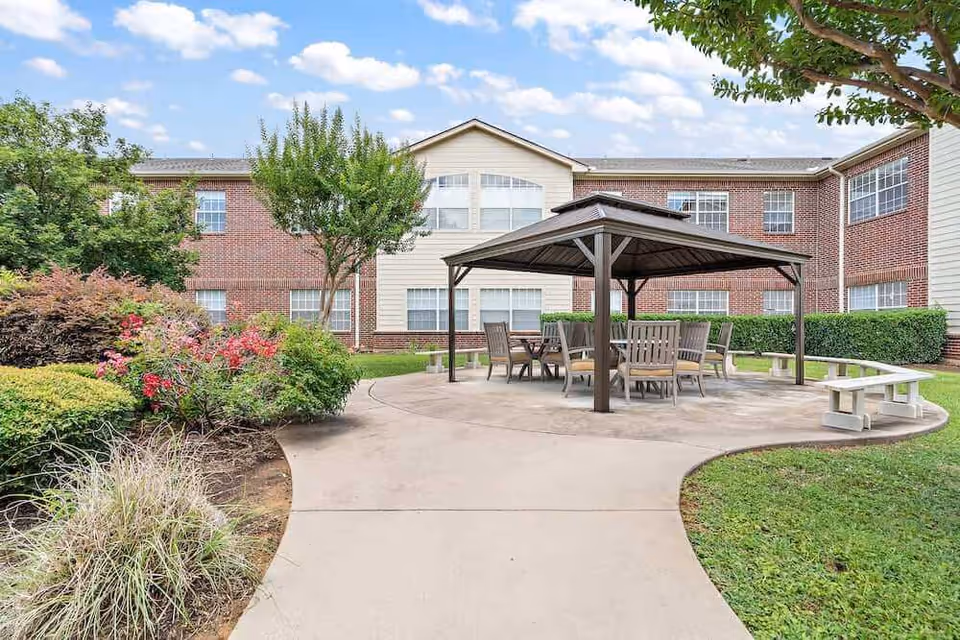 Outdoor patio area at Morada Lake Arlington with a paved walkway leading to a covered seating area featuring a table and several chairs. The patio is surrounded by green grass, bushes, and trees, with a two-story brick and siding building in the background under a partly cloudy sky.