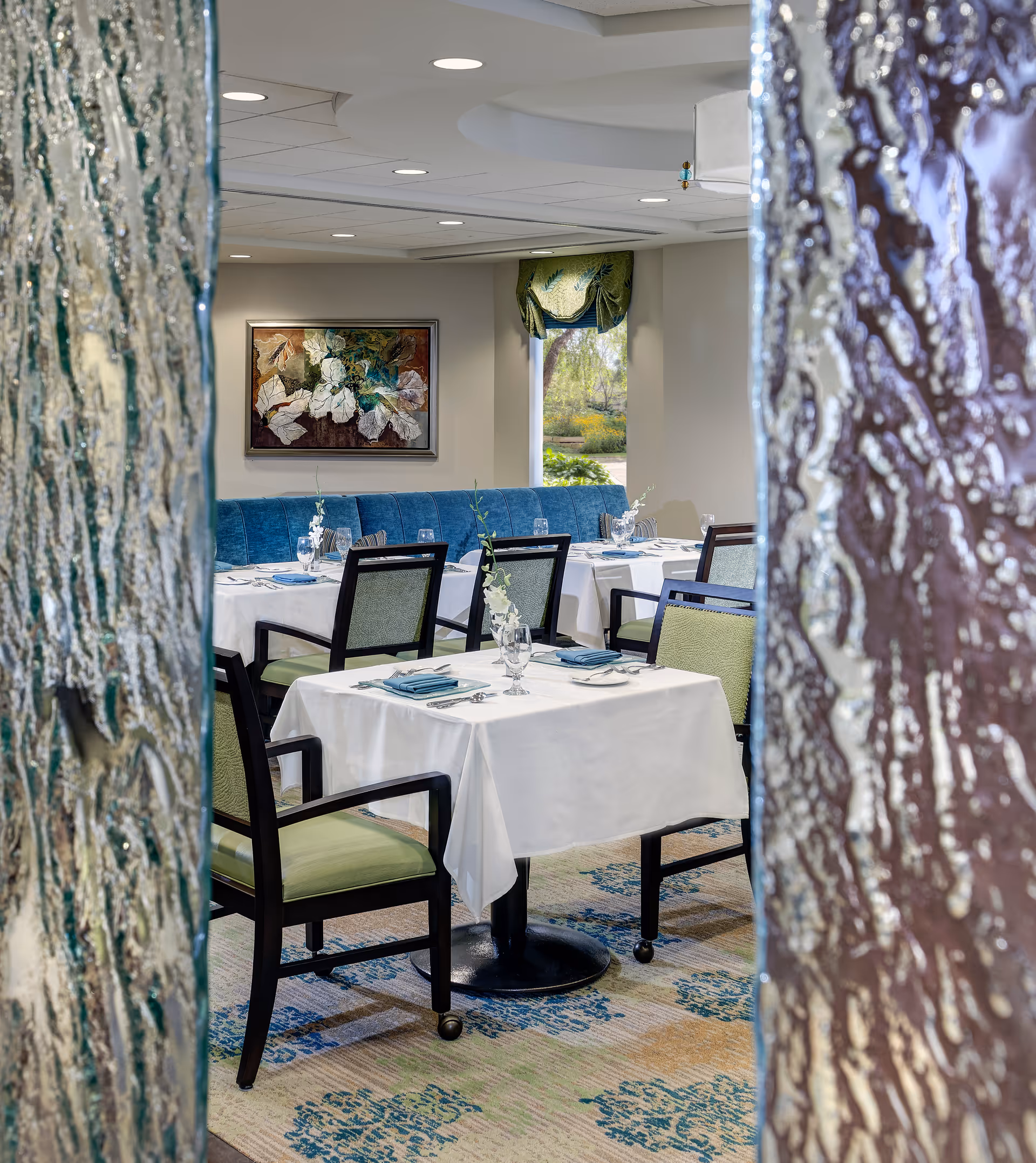 Dining room with tables set with white tablecloths, green-upholstered chairs and a blue banquette, viewed between textured glass panels.