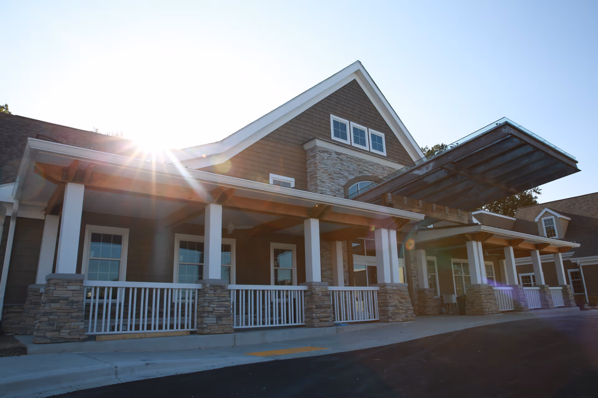 Exterior view of a senior living facility building with stone and wood siding, white pillars, and a covered entrance. The sun is shining brightly in the background.