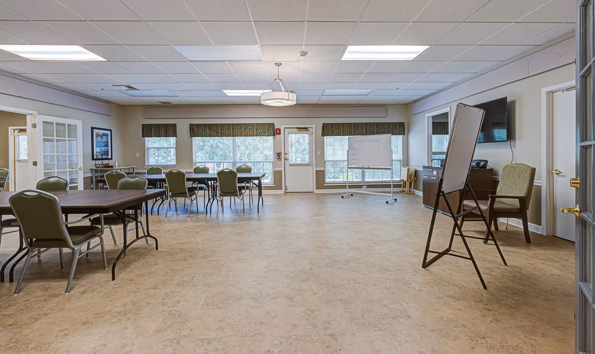 A spacious, well-lit room with multiple tables and chairs arranged for group activities or dining. The room has large windows with valances, a whiteboard on a stand, a TV mounted on the wall, and a few armchairs. The floor is covered with light-colored tiles, and the ceiling has recessed lighting and a hanging light fixture.