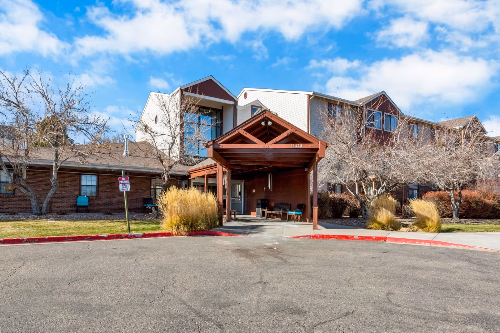 Front exterior view of Northglenn Heights Assisted Living and Memory Care building with a covered entrance, leafless trees, ornamental grasses, and a clear blue sky with some clouds.