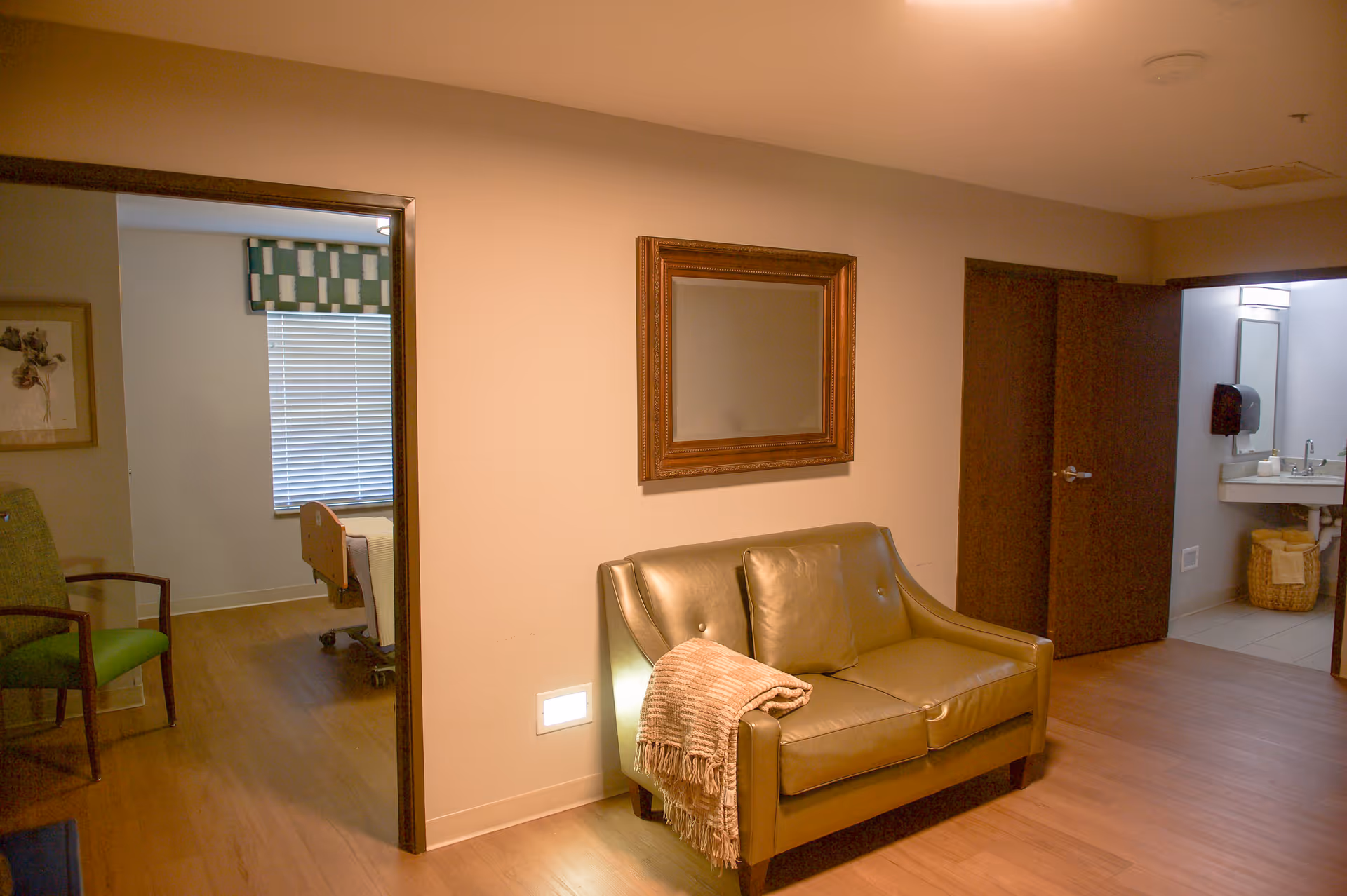 Interior view of a senior living facility room with a green leather loveseat with a throw blanket and pillow, a large framed mirror on the wall above it, an open doorway leading to a bedroom with a hospital-style bed and a window with blinds, and another open doorway leading to a bathroom with a sink and mirror.