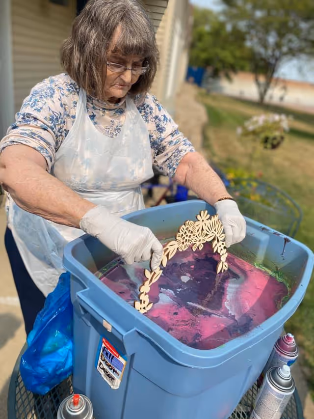 An elderly woman wearing glasses, a floral shirt, a white apron, and gloves is holding a wooden decorative piece with flower shapes over a large blue plastic bin filled with colorful water outdoors. There are spray paint cans on the table next to the bin, and a grassy area with trees is visible in the background.
