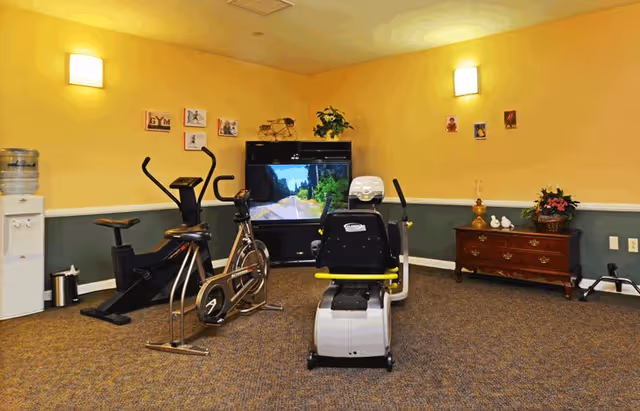A small exercise room with three stationary exercise bikes facing a large television screen displaying a road scene. The room has yellow walls with green wainscoting, carpeted floor, wall-mounted lights, a water cooler, and a wooden dresser with decorative items and plants.