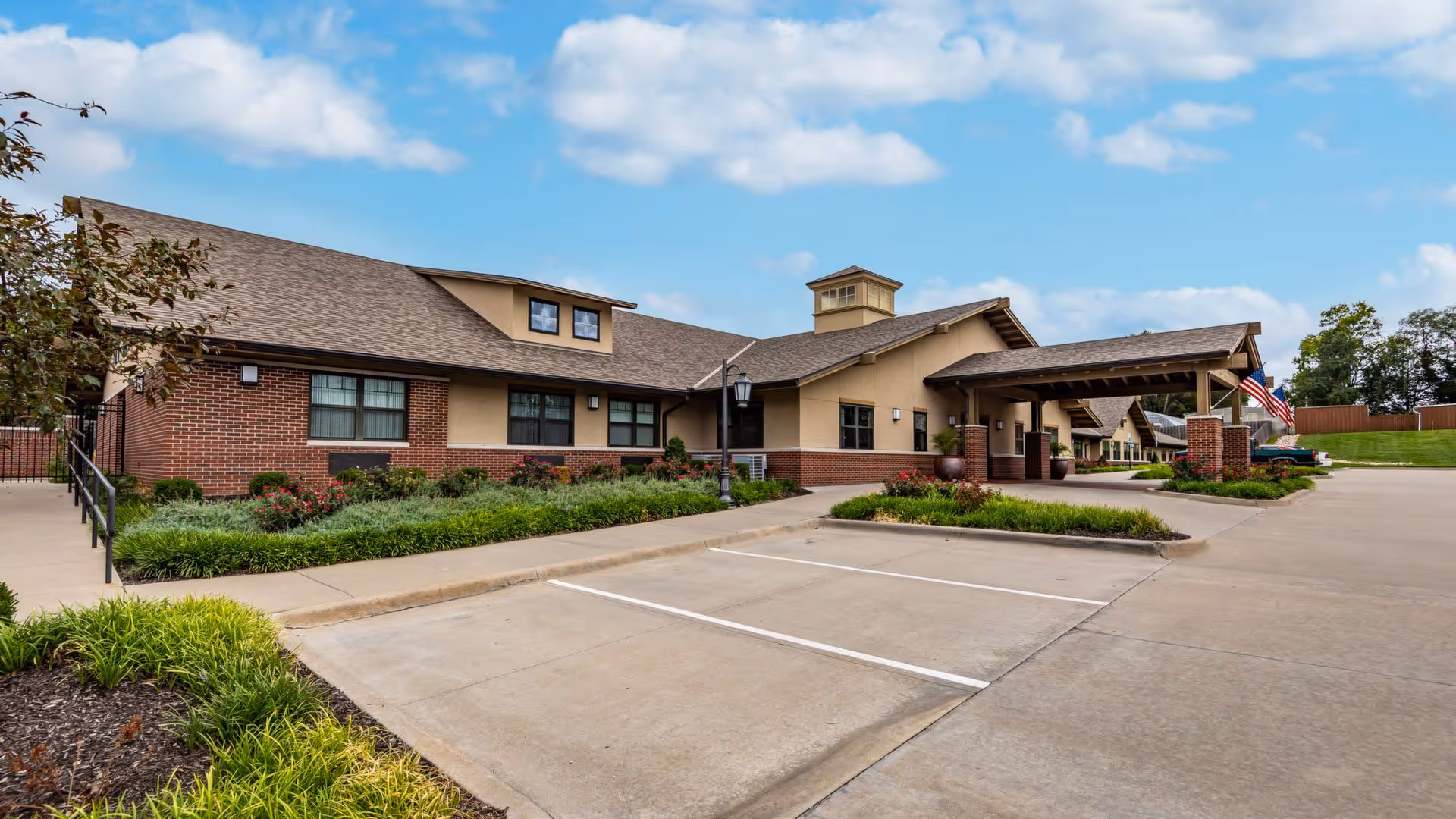 Exterior view of a single-story senior living facility building with a covered entrance, brick and beige siding, landscaped greenery, and a clear blue sky with some clouds.