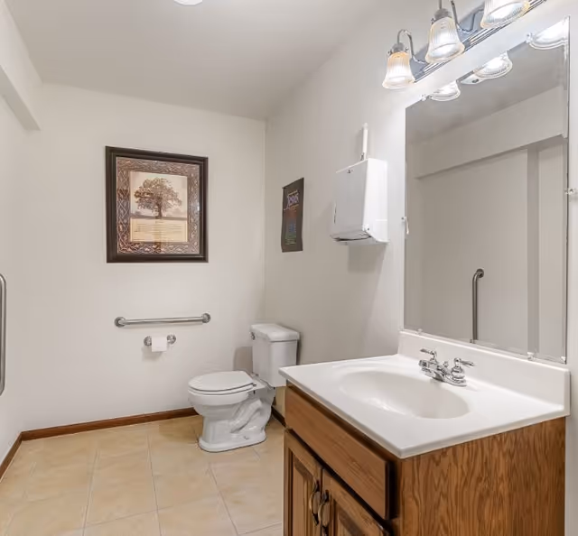 A clean bathroom with a white toilet, a wooden vanity with a white sink, a large mirror above the sink, a wall-mounted paper towel dispenser, and a framed picture on the wall. The floor is tiled and there is a grab bar next to the toilet.