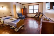 A nursing facility patient room with a hospital bed, armchairs, dresser with a TV, and a window over wood floors.
