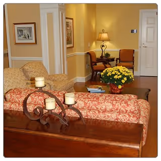 A cozy living room area with a patterned red sofa, a wooden table holding three lit candles, a beige armchair, a coffee table with a yellow flower arrangement and books, and two orange chairs against a yellow wall with framed artwork and a lamp.