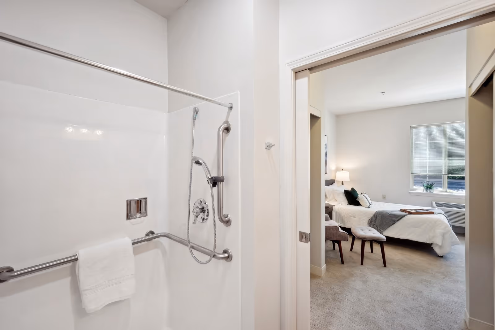 View of a bathroom with a white shower area equipped with grab bars and a handheld showerhead, a white towel hanging on a bar, and an adjacent bedroom with a bed, nightstand with lamp, chair, and window with blinds.
