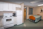 A rehab-style room showing a small kitchenette with stove, refrigerator and cabinets and a single adjustable bed with green bedding.