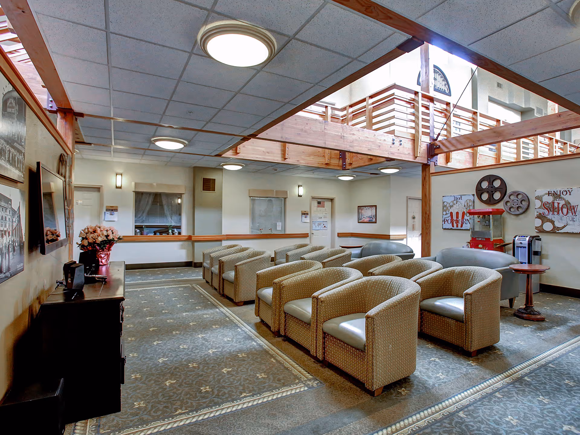 A cozy senior living common area with rows of cushioned armchairs facing a wall-mounted TV. The room features a popcorn machine, movie-themed wall decorations, and a coffee maker on a small round table. The ceiling has recessed lighting and wooden beams, with a second-floor balcony visible above.
