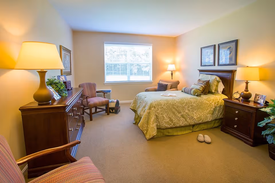 A cozy bedroom with a large window letting in natural light. The room features a bed with green patterned bedding, multiple pillows, and a book placed on it. There are two wooden nightstands on either side of the bed, each with a lamp and decorative items. A wooden dresser with a lamp, framed photos, and a plant is on the left side of the room. Two upholstered chairs are positioned near the dresser and window. The walls are painted a soft beige color, and the carpet is light brown.