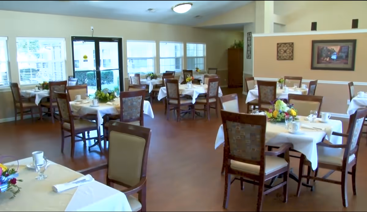 A dining room in a senior living facility with multiple tables covered in white tablecloths, each set with cups, glasses, napkins, and floral centerpieces. The room has wooden chairs with cushioned seats and backs, large windows letting in natural light, and framed artwork on the walls.