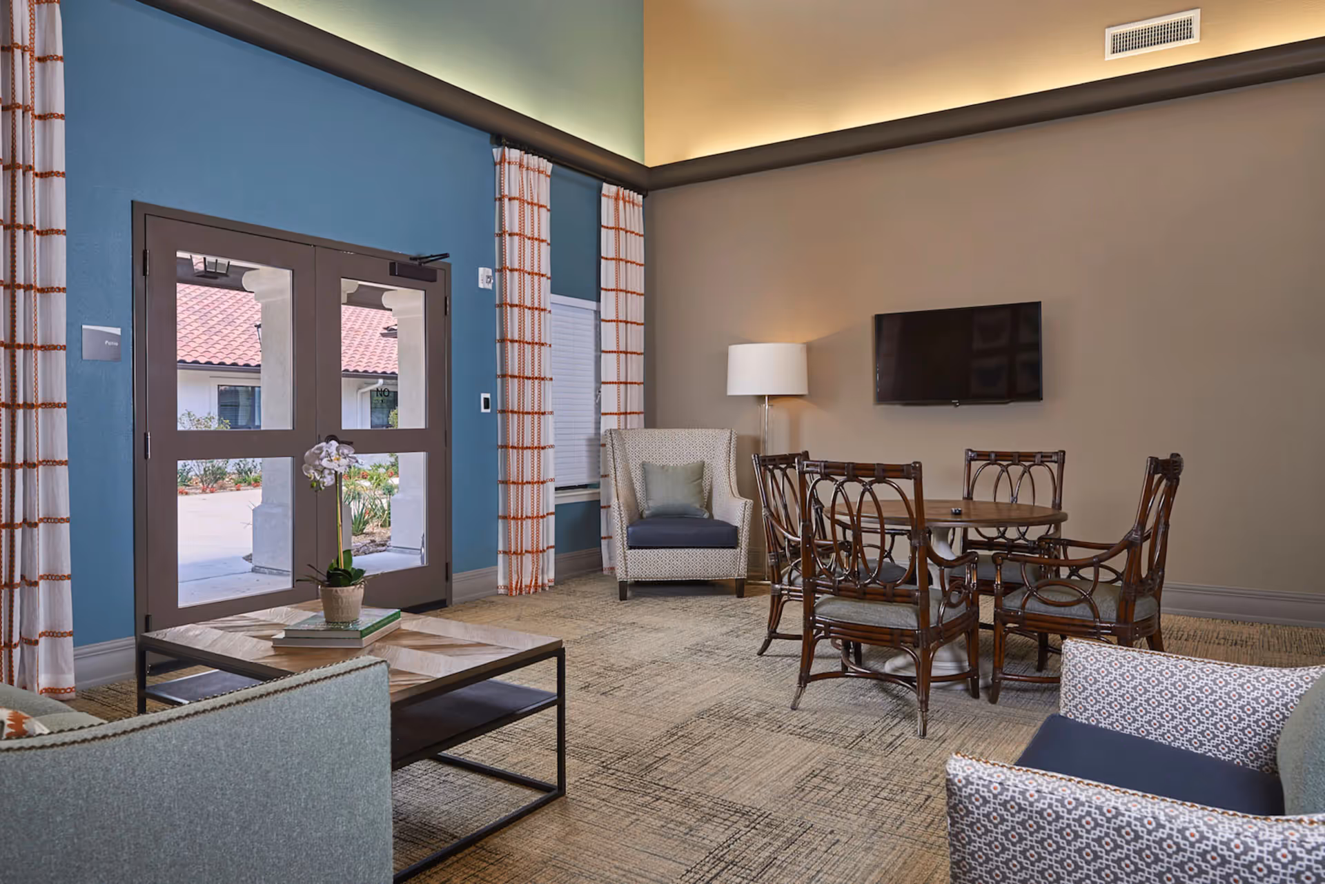 A bright communal living room with armchairs, a coffee table, a round dining table with chairs, a wall-mounted TV and glass double doors.