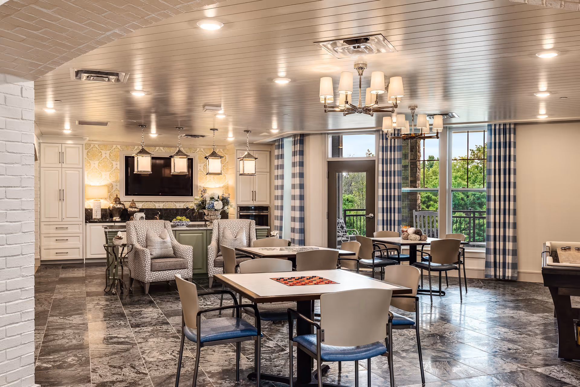 A spacious, well-lit common area with multiple tables and chairs arranged for social activities. The room features a kitchen area with white cabinetry, a large TV mounted on the wall, and decorative lighting fixtures. Large windows with blue and white checkered curtains allow natural light to fill the space, and there is a door leading to an outdoor area. The floor is tiled with a dark marble pattern, and the ceiling has recessed lighting and chandeliers.