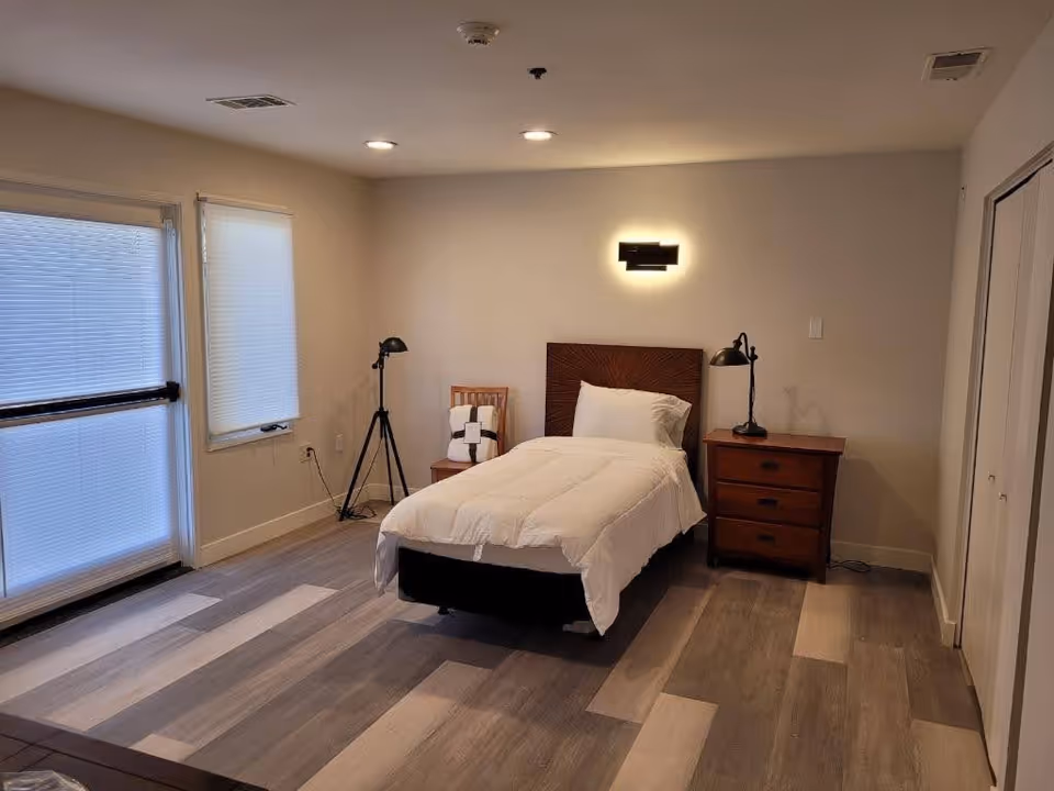 A simple bedroom with a single bed covered in white bedding, a wooden headboard, and a pillow. There is a wooden nightstand with a black lamp on the right side of the bed and a wooden chair with a folded blanket on it to the left. The room has light-colored walls, a window with closed blinds, and a door with blinds on the left side. The floor is covered with light and dark wood-patterned vinyl or laminate planks. Ceiling lights and a wall-mounted light fixture above the bed provide illumination.