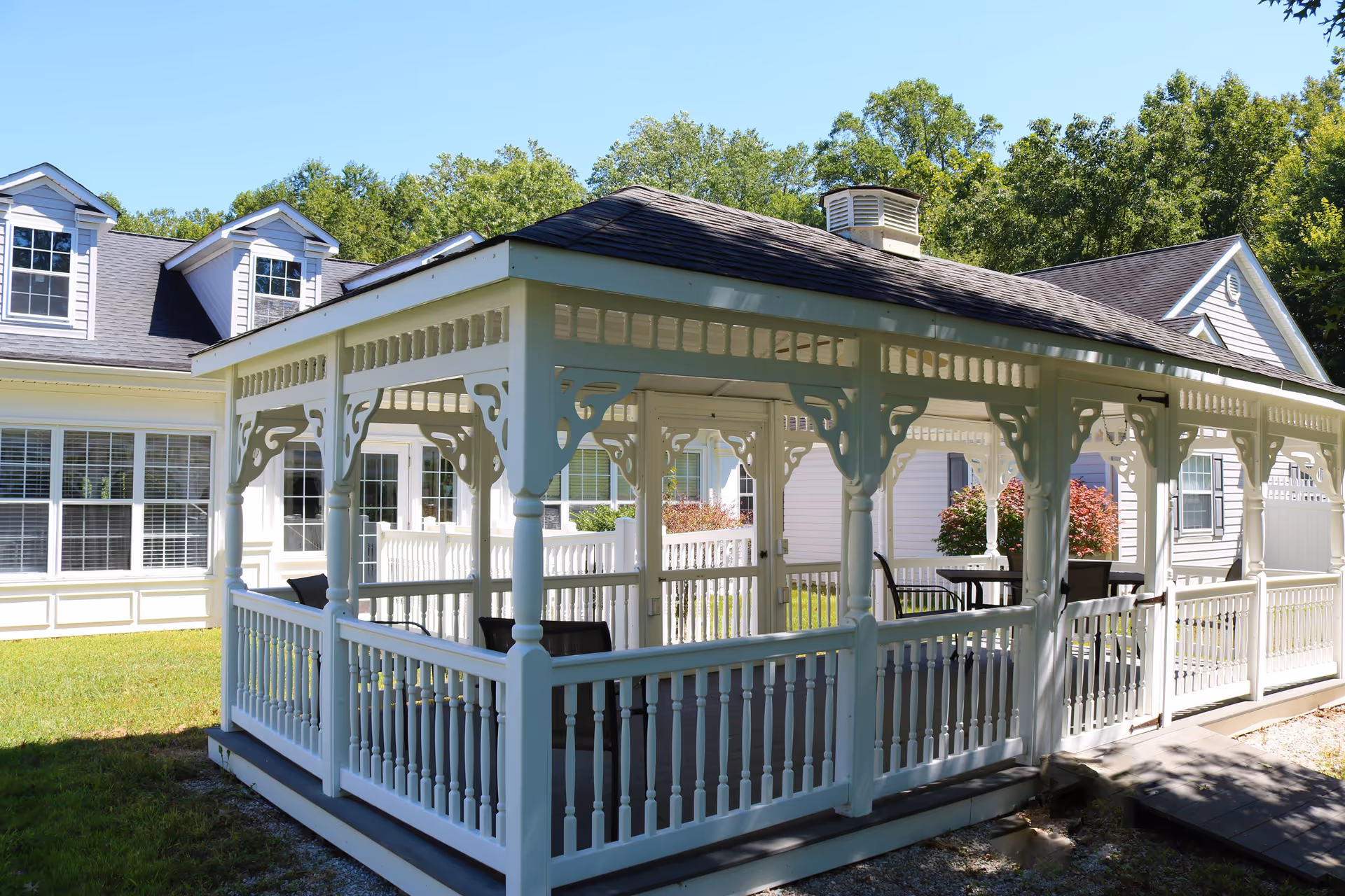 A white wooden gazebo with decorative trim and railings situated outdoors on a grassy area, with a building featuring large windows and dormer windows in the background, surrounded by trees and shrubs.