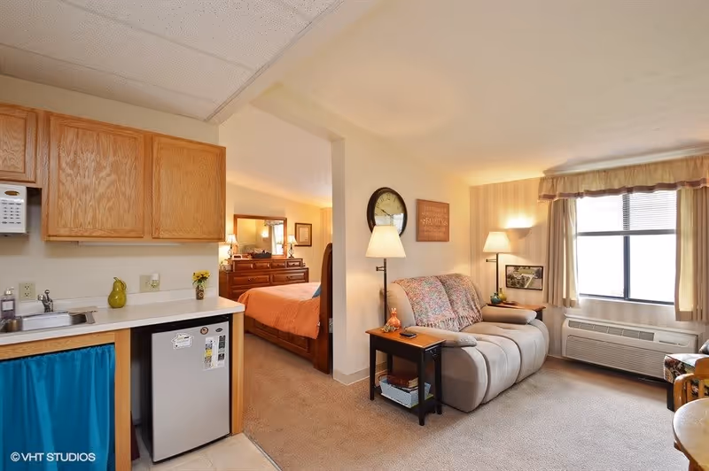 Interior view of a senior living facility room showing a small kitchenette with wooden cabinets, a mini fridge, and a sink on the left. In the center, there is a cozy living area with a beige recliner sofa, a side table with a lamp, and a wall clock. To the right, a window with curtains lets in natural light. In the background, a bedroom with a wooden bed frame and dresser is visible.