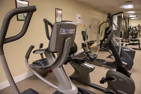 Interior view of a fitness room in Glen Meadows Retirement Community featuring several exercise machines including stationary bikes and elliptical trainers, with beige walls and framed pictures hanging on the wall.