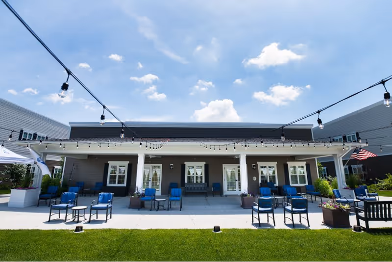 Outdoor covered patio with blue chairs, tables and string lights in front of a senior living building.