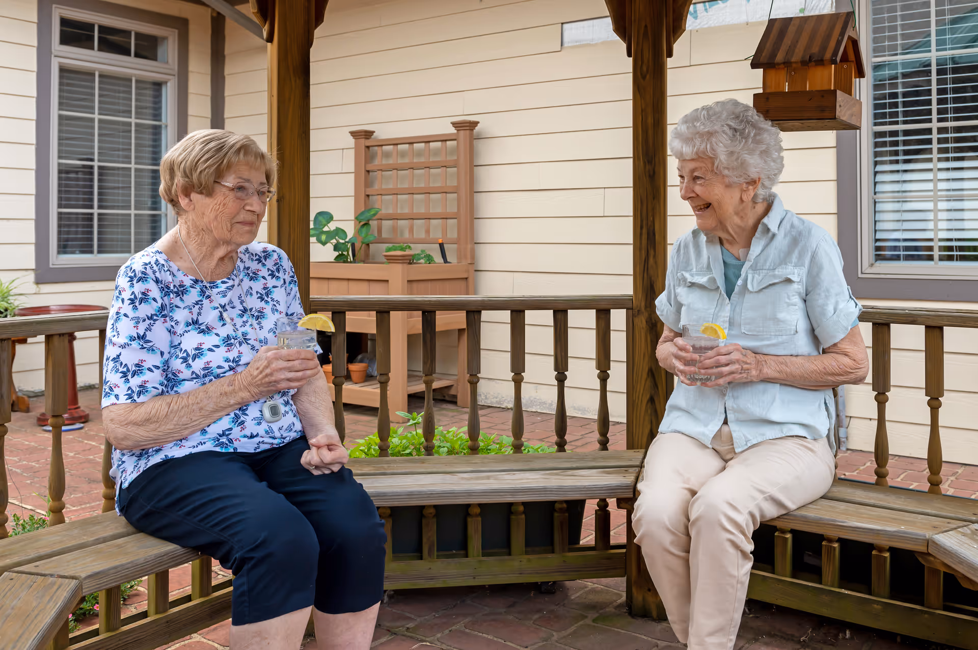 Two elderly women sitting on a wooden bench under a gazebo, each holding a glass of water with a lemon slice, smiling and engaging in conversation. The setting is an outdoor patio area with beige siding walls, windows, and some potted plants in the background.