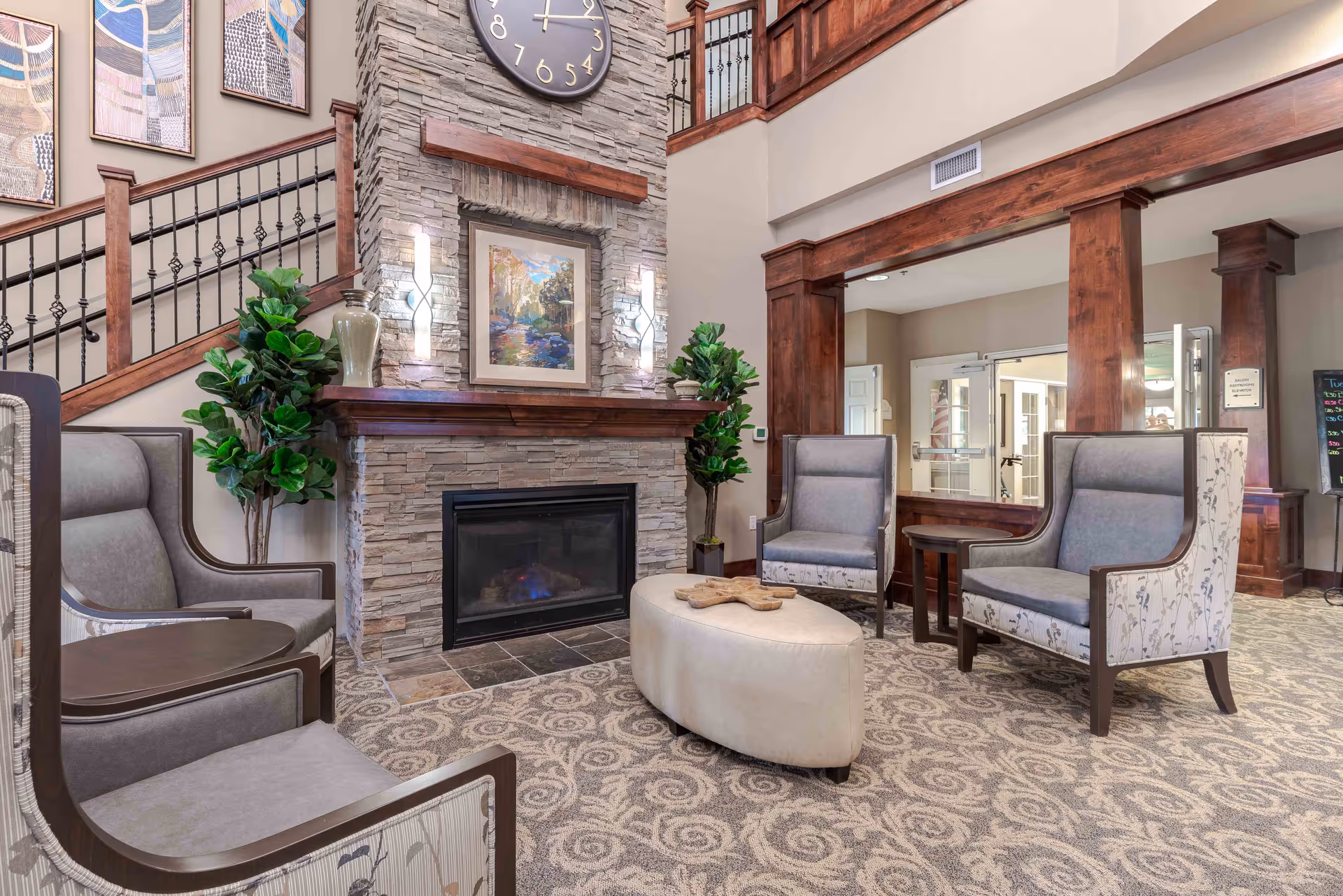 A cozy and elegant common area in a retirement community featuring a stone fireplace with a large clock above it, surrounded by four upholstered armchairs and a cushioned ottoman in the center. The room has wooden accents, decorative plants, patterned carpet, and a staircase with wooden railings in the background.