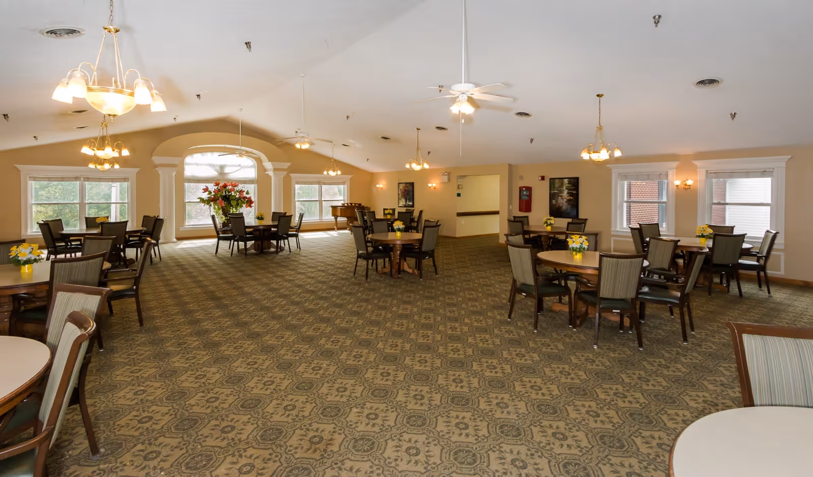 A spacious dining room with multiple round tables and chairs arranged neatly. The room features large windows allowing natural light, ceiling fans, chandeliers, and floral centerpieces on the tables. The carpet has a patterned design, and there is a piano in the background near the windows.
