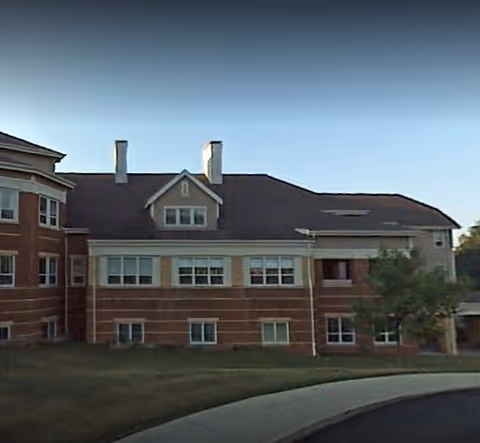 Exterior view of a multi-story brick building with multiple windows and a sloped roof under a clear sky, surrounded by a grassy area and a curved driveway.