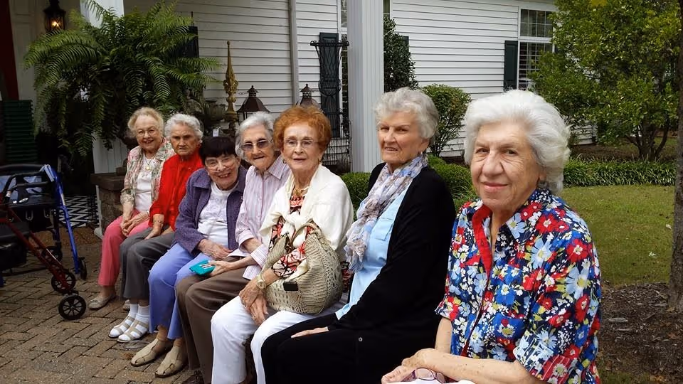 Seven elderly women sitting on a bench outside a white building with greenery around them. They are smiling and appear to be enjoying each other's company on a paved area near the entrance.