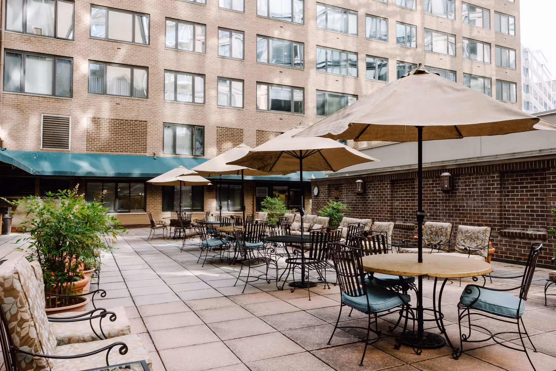 Outdoor patio area at Thomas House Retirement Community with multiple round tables, metal chairs with cushions, large beige umbrellas, potted plants, and a brick building in the background.