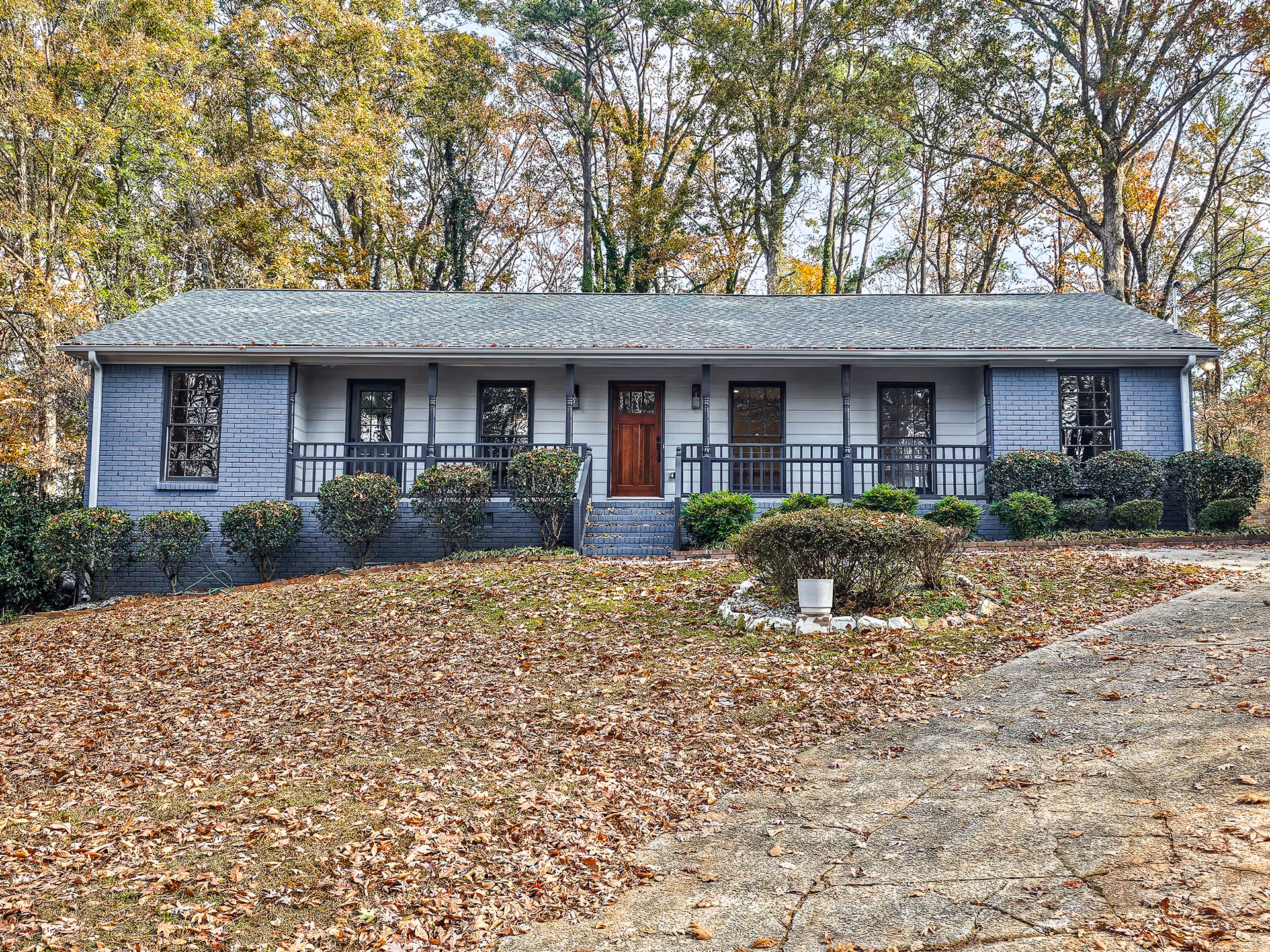 Front exterior view of a single-story assisted living facility building with a gray roof, blue-gray brick and siding walls, a wooden front door, and a porch with railings. The building is surrounded by bushes and trees with autumn foliage, and there is a driveway and a small circular garden with a white planter in front.