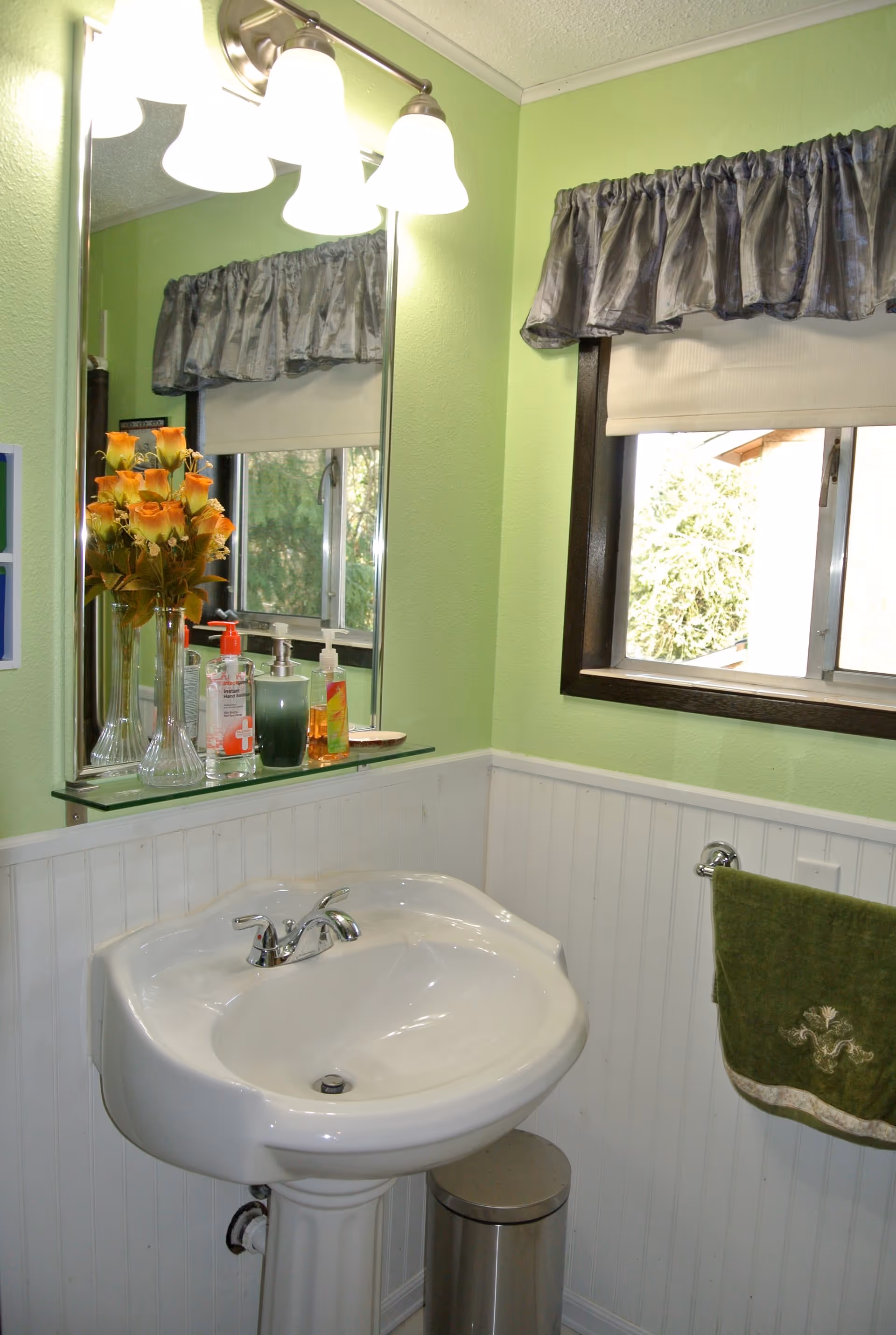 A bathroom corner with a white pedestal sink, a mirror above it with three light fixtures, a glass shelf holding a vase of orange roses and various soap dispensers. There is a window with a gray valance and a white blind, and a green towel hanging on a towel ring on the wall. The walls are painted light green with white wainscoting, and a small silver trash can is placed under the sink.