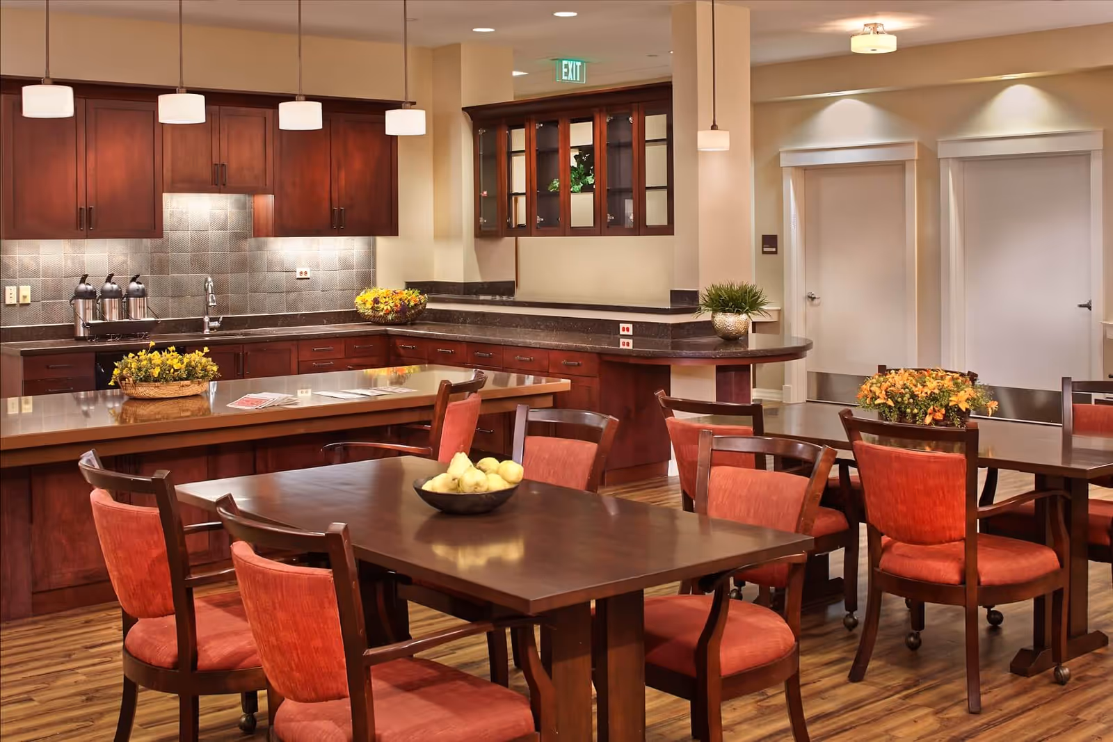 A warm and inviting kitchen and dining area with wooden cabinets, a tiled backsplash, and multiple pendant lights hanging from the ceiling. There are several wooden tables with red cushioned chairs, decorated with bowls of fruit and flower arrangements. The floor is wooden, and there are two white doors in the background.