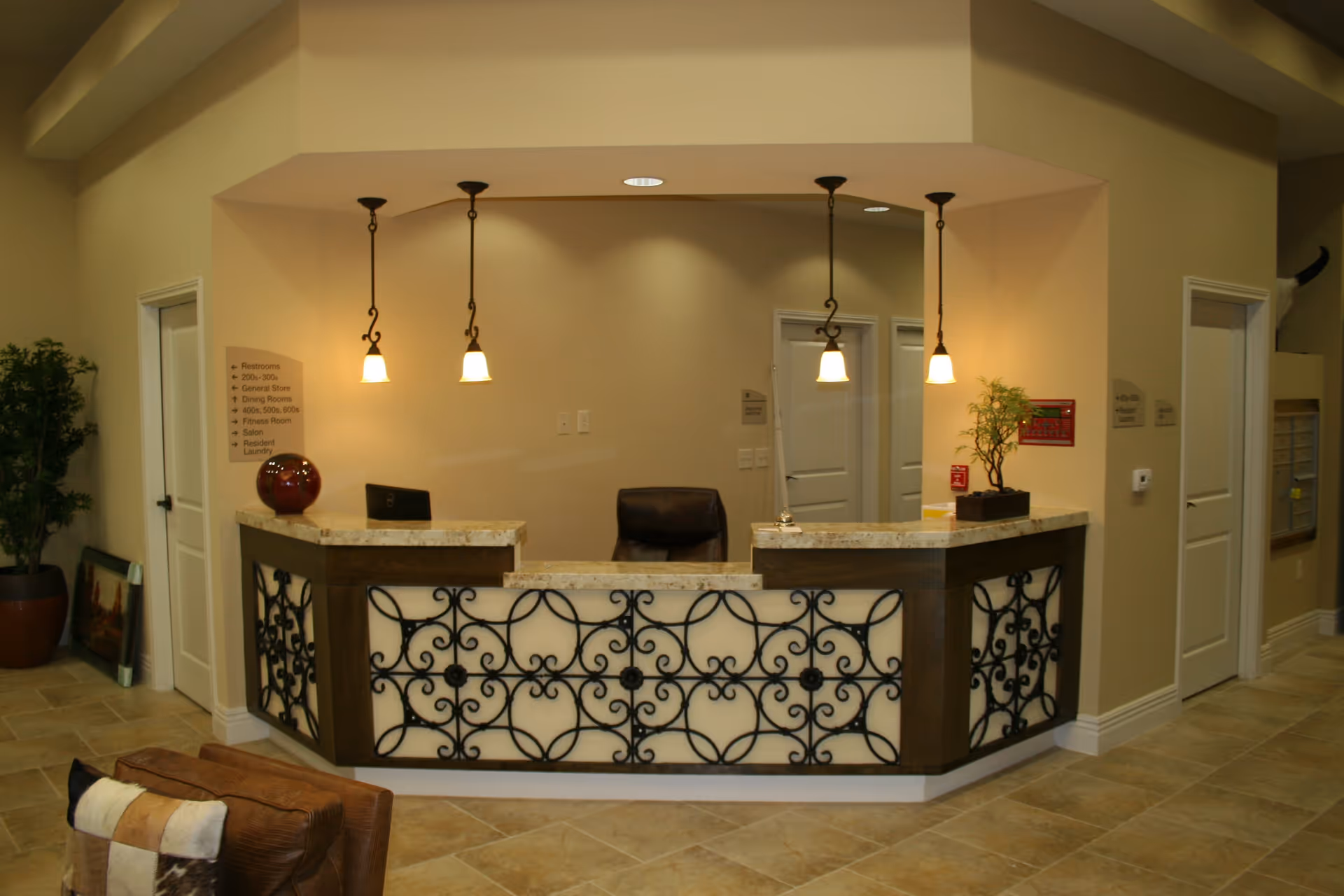 Reception desk area with decorative wrought iron panels and three hanging pendant lights. Behind the desk is a black office chair and beige walls with doors on either side. There is a potted plant on the left and a small plant on the right side of the desk. The floor is tiled in a light beige color.