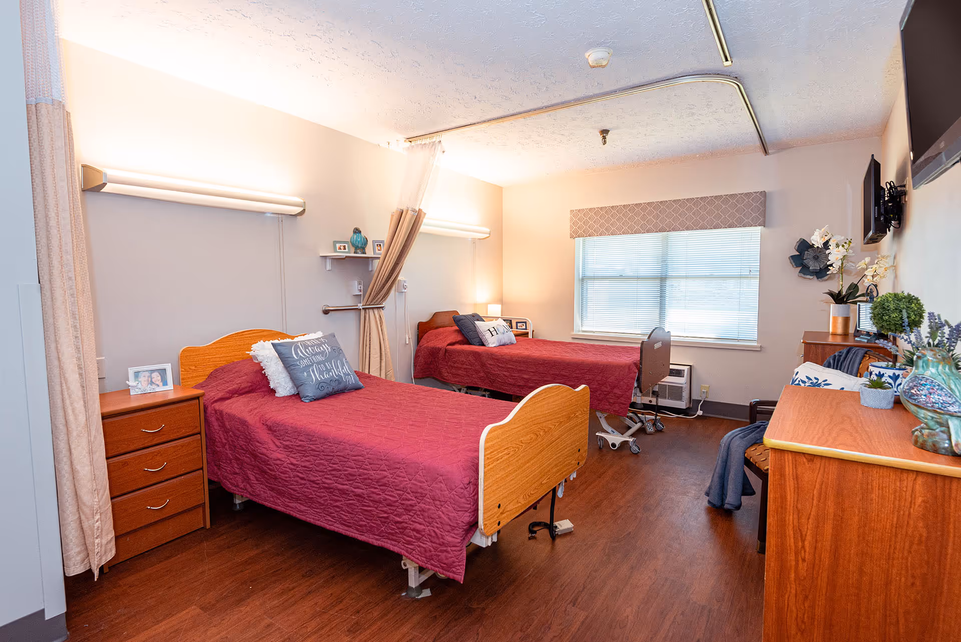 A well-lit room with two single beds covered in red quilts, each with decorative pillows. There is a wooden nightstand with drawers between the beds, a window with blinds and a valance, and a TV mounted on the wall. The room has wooden flooring and is decorated with plants and framed photos.