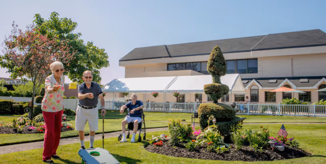 Three elderly individuals enjoying an outdoor garden area at an assisted living facility. One woman is tossing a bean bag towards a cornhole board, a man with a cane is standing nearby watching, and another man is seated in a wheelchair. The background shows a large building with a white awning and well-maintained landscaping with flowers and trimmed bushes.