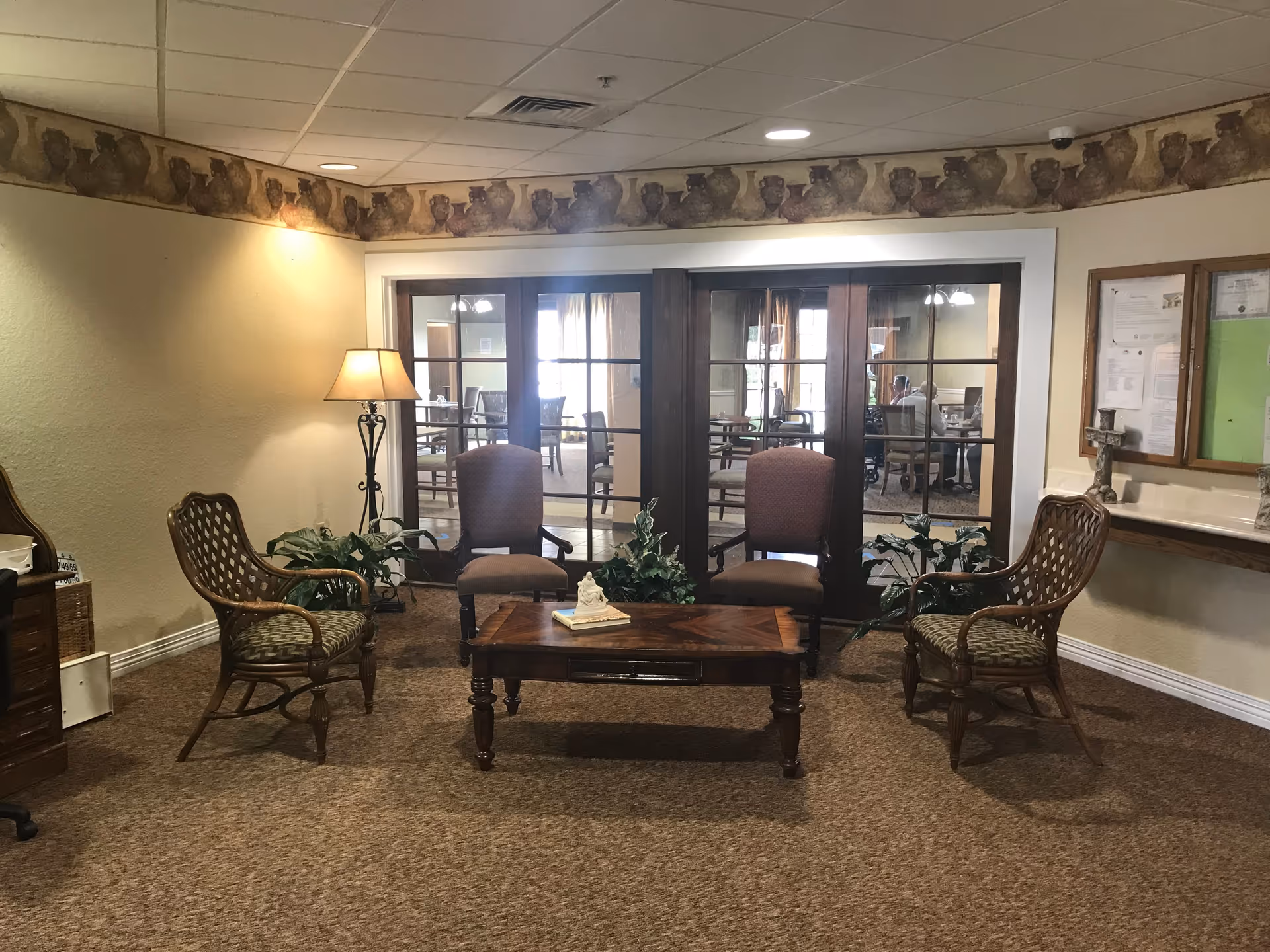 A cozy seating area in a senior living facility with four chairs arranged around a wooden coffee table. The room has beige walls with a decorative border near the ceiling, a floor lamp, and some potted plants. Behind the seating area are glass-paneled double doors leading to another room with more chairs and tables.
