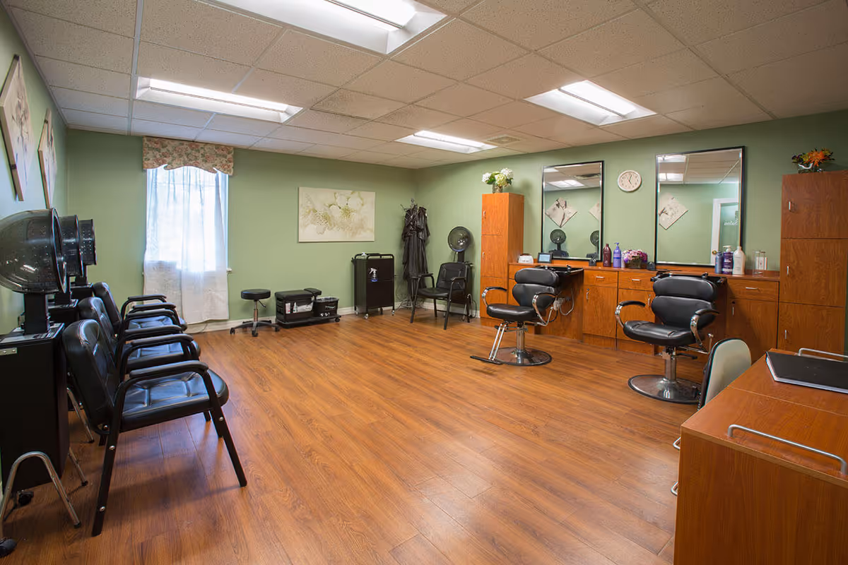 Interior of a hair salon room with green walls and wooden flooring. The room features two black salon chairs in front of a wooden counter with mirrors, hair care products, and a clock on the wall. On the left side, there are several black chairs with hair dryers above them, and a window with a floral valance and white curtain. Additional salon equipment and storage cabinets are also visible.