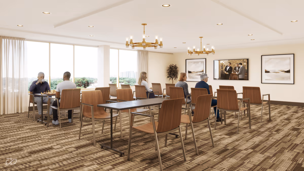A spacious, well-lit common room with large windows showing a view of greenery outside. Several people are seated on wooden chairs arranged in rows and at tables. Two individuals are playing chess near the windows, while others are seated facing a television mounted on the wall displaying a scene with three men in suits. The room features beige walls, patterned carpet, modern chandeliers, and framed artwork on the walls.