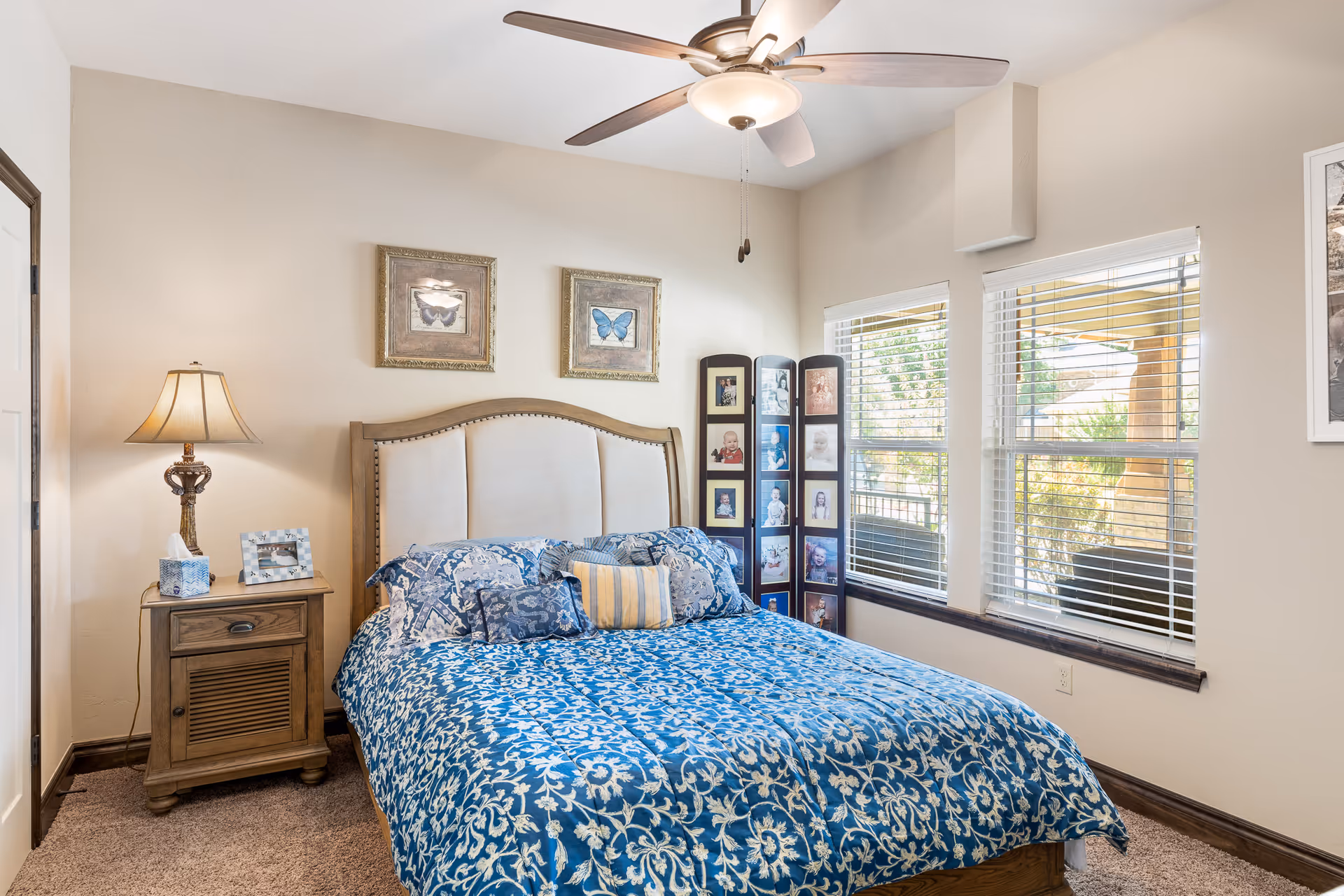Well-lit bedroom with a blue patterned bedspread, upholstered headboard, nightstand with a lamp, framed art, and windows with blinds.
