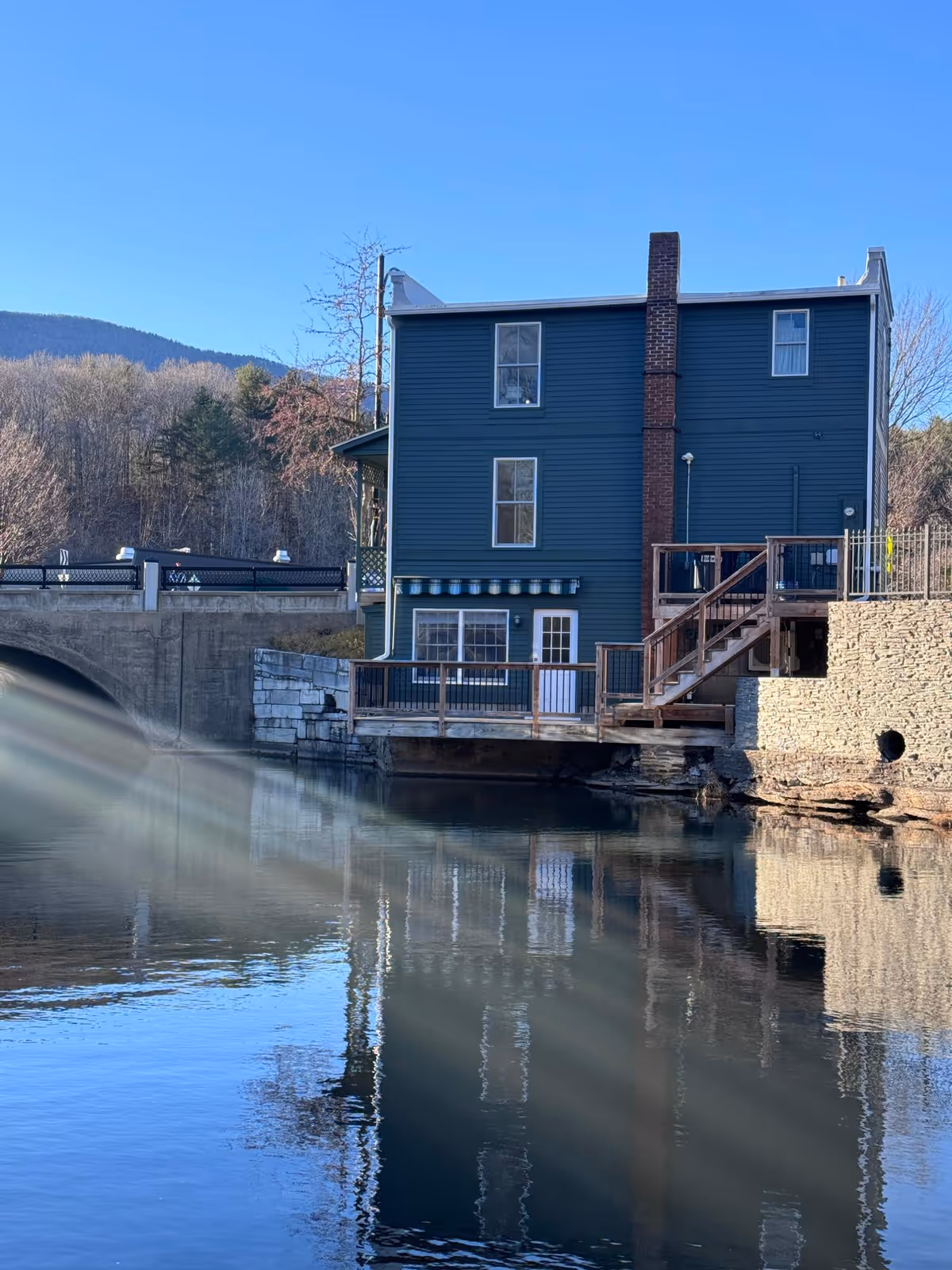 Blue three-story riverside building with a wooden deck and stairs reflected in calm water under a clear blue sky.