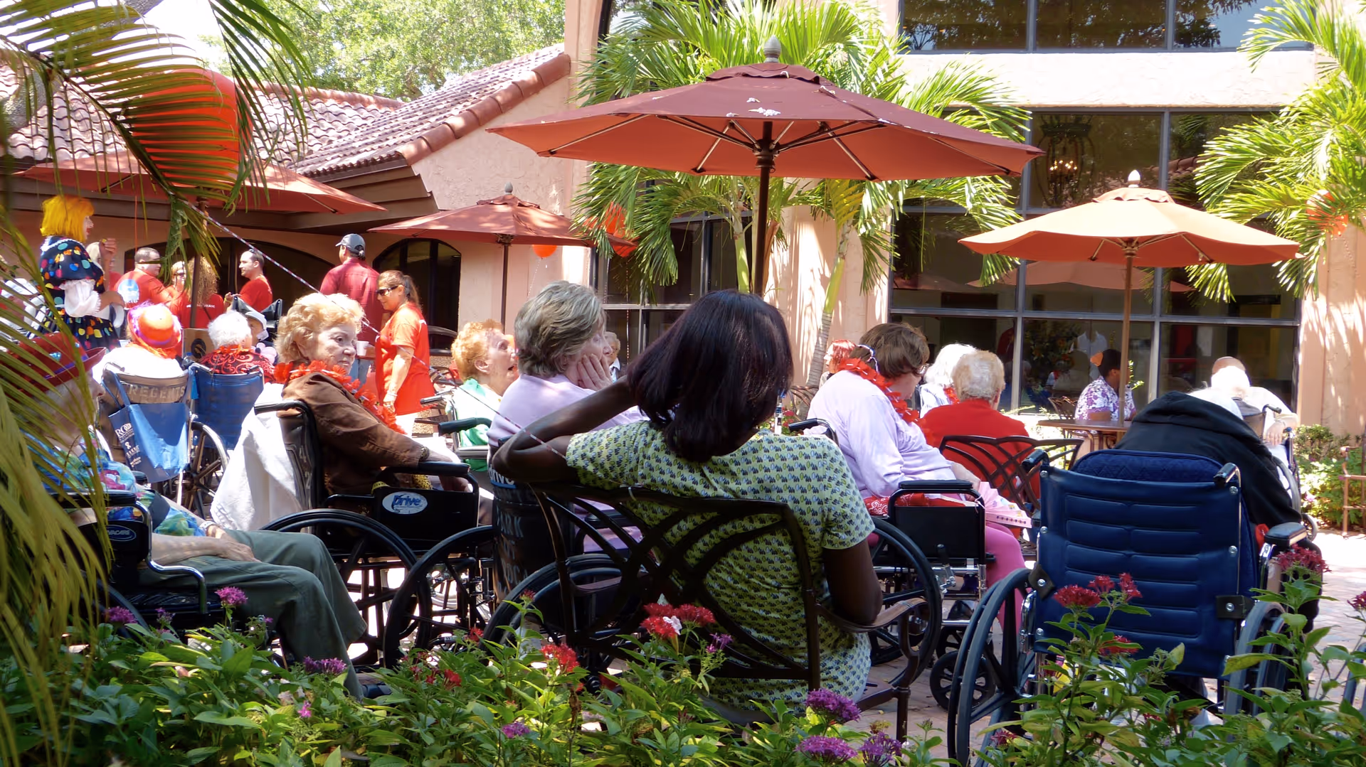 A group of elderly people, many in wheelchairs, sitting outdoors under red umbrellas in a garden courtyard with palm trees and flowering plants. Several caregivers or staff members are standing and interacting with the residents. The building with large windows is visible in the background.