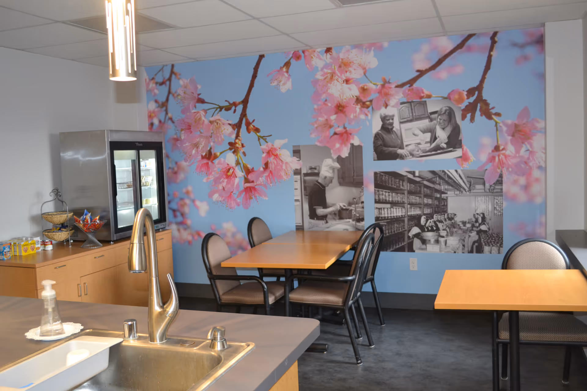 A small dining area with wooden tables and cushioned chairs, a stainless steel sink and faucet in the foreground, a refrigerated display case on a wooden cabinet, and a wall decorated with a large mural of pink cherry blossoms and three black-and-white historical photos depicting people cooking and working in a kitchen.