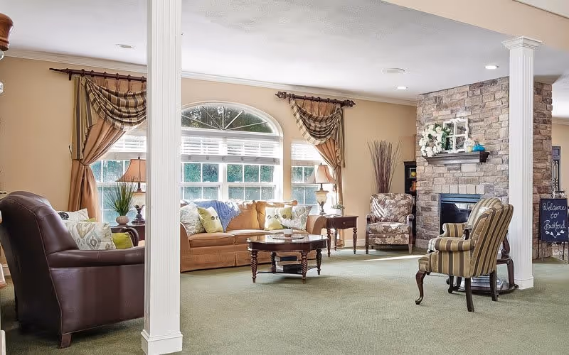 A spacious and well-lit living room area in a senior living facility featuring a large window with curtains, a brown leather armchair, a beige sofa with colorful cushions, a wooden coffee table, two patterned armchairs, a stone fireplace decorated with flowers, and a green carpeted floor.
