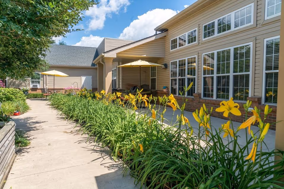 Outdoor patio area at Addington Place of Northville with a concrete walkway lined by yellow flowers and greenery. The building exterior features large windows and beige siding. There are patio tables with yellow umbrellas and seating areas under a covered section. The sky is blue with some clouds.
